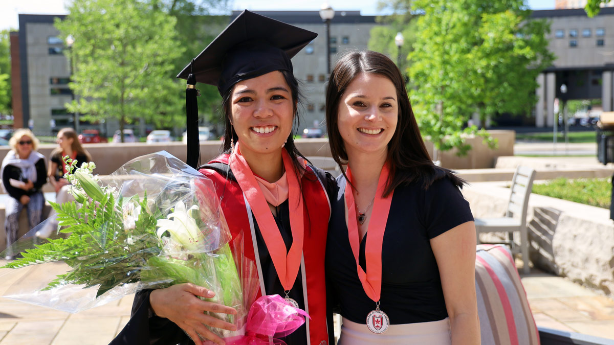 Two new public health graduates, one holding flowers and wearing commencement regalia 
