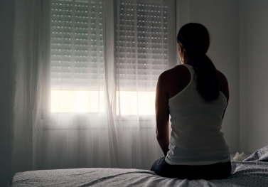 A woman sits on a bed looking toward a window