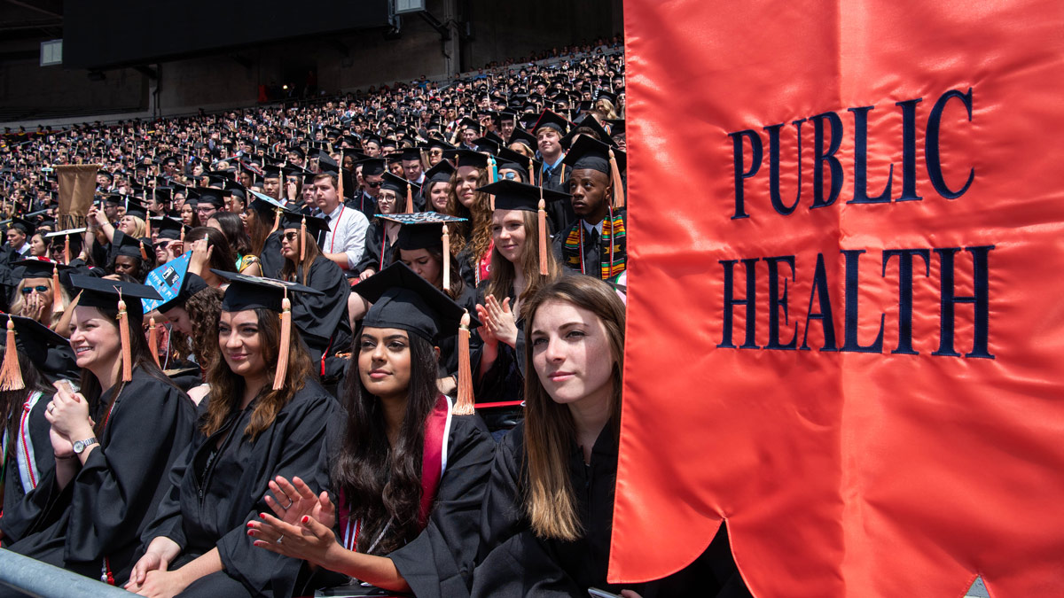 Public Health students in caps and gowns at graduation in Ohio Stadium. A salmon-colored banner with the words "public health" fills the right side of the image.