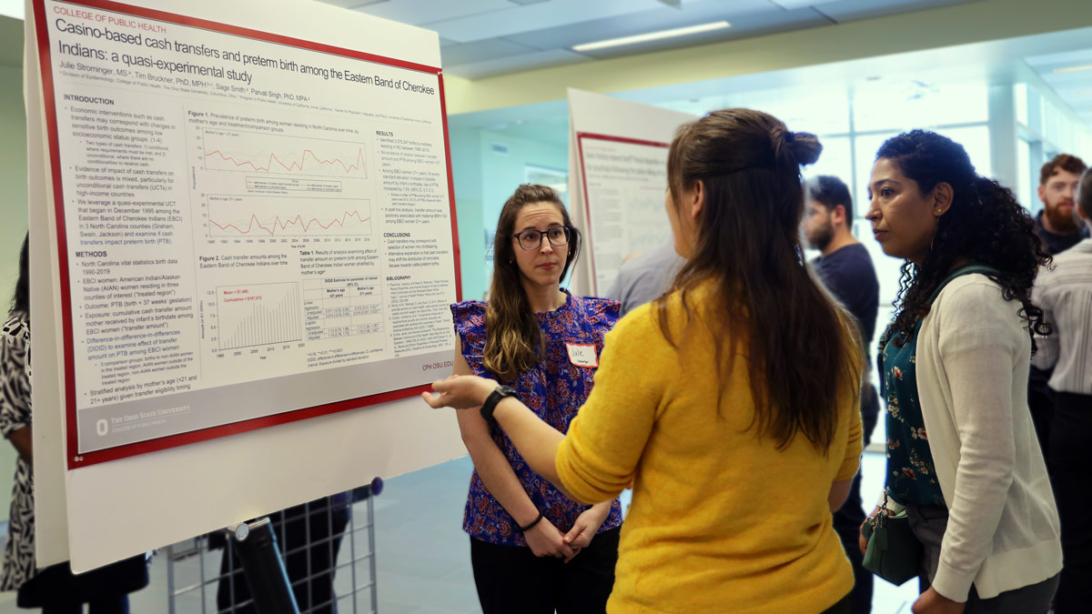 Three women stand discussing epidemiology research at a poster presentation