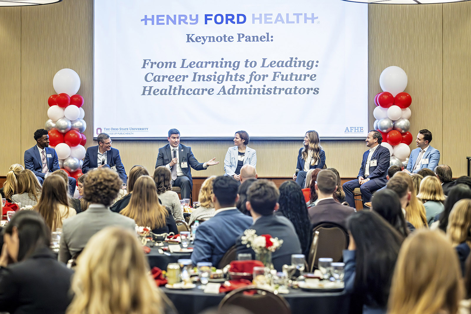 People dressed in business attire speak in front of a crowded room.