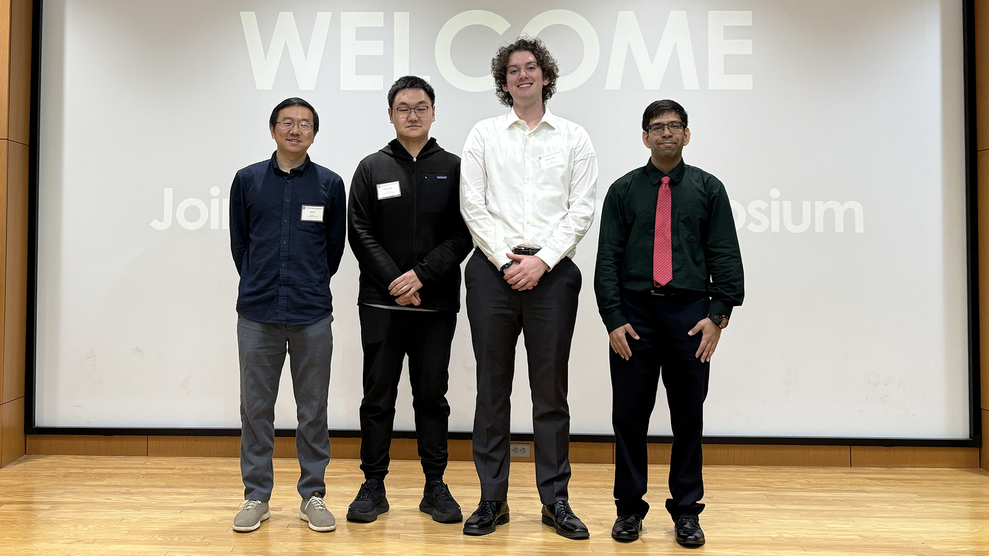 Four people stand in a row smiling with a sign that says "welcome" in the background.