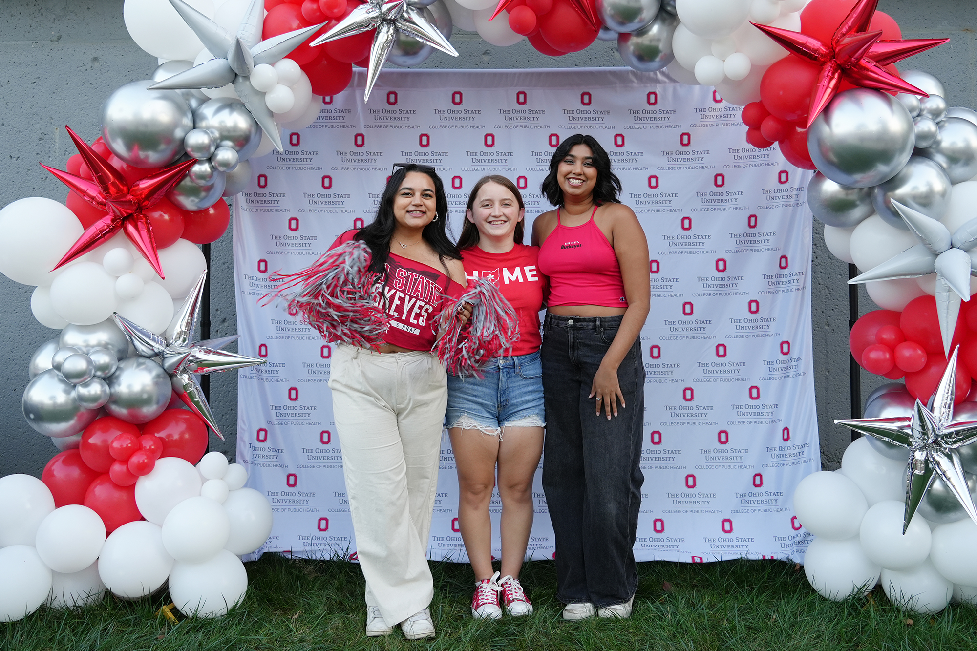 three people in Buckeye spirit gear stand in front of an Ohio State background with balloons