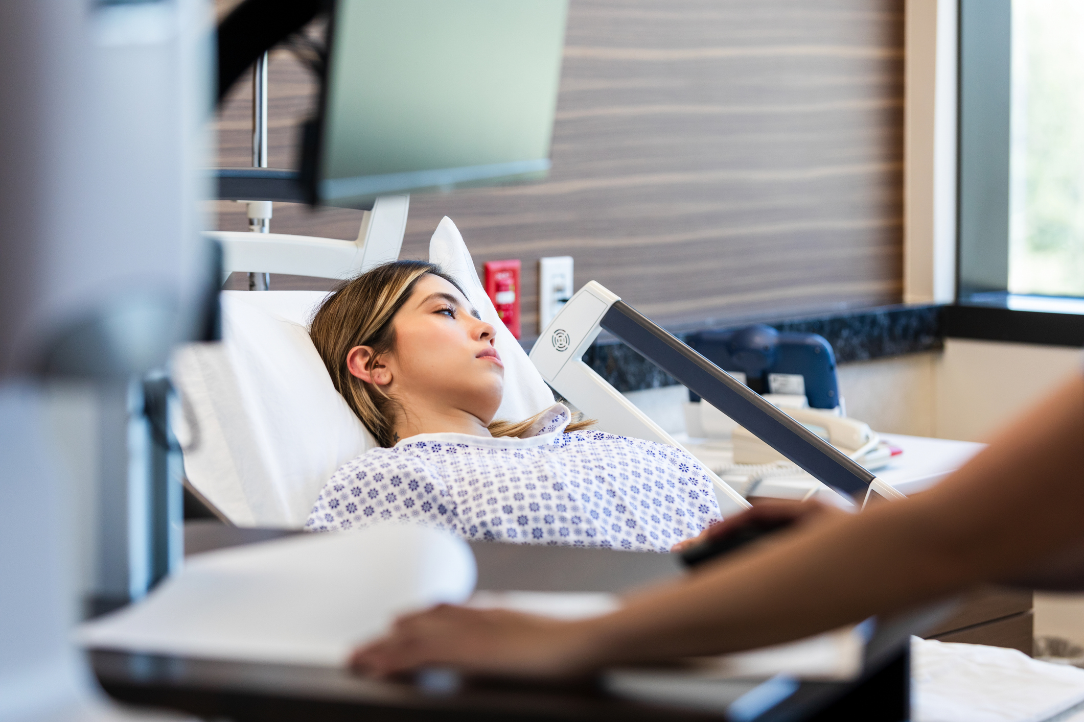 A young woman in a hospital bed with a health care professional's arm in the foreground
