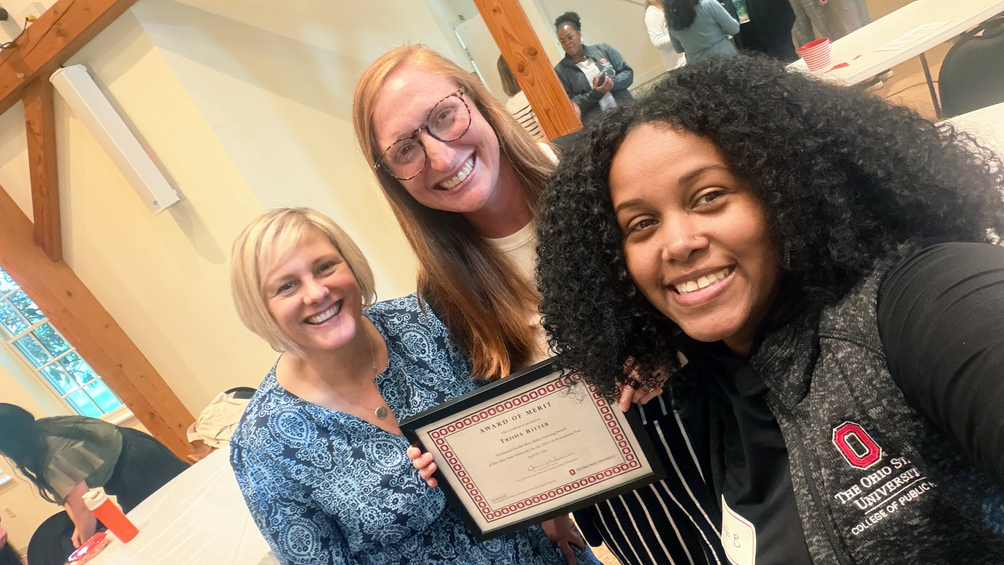 Three people smile indoors. One person holds a certificate that says "award of merit."