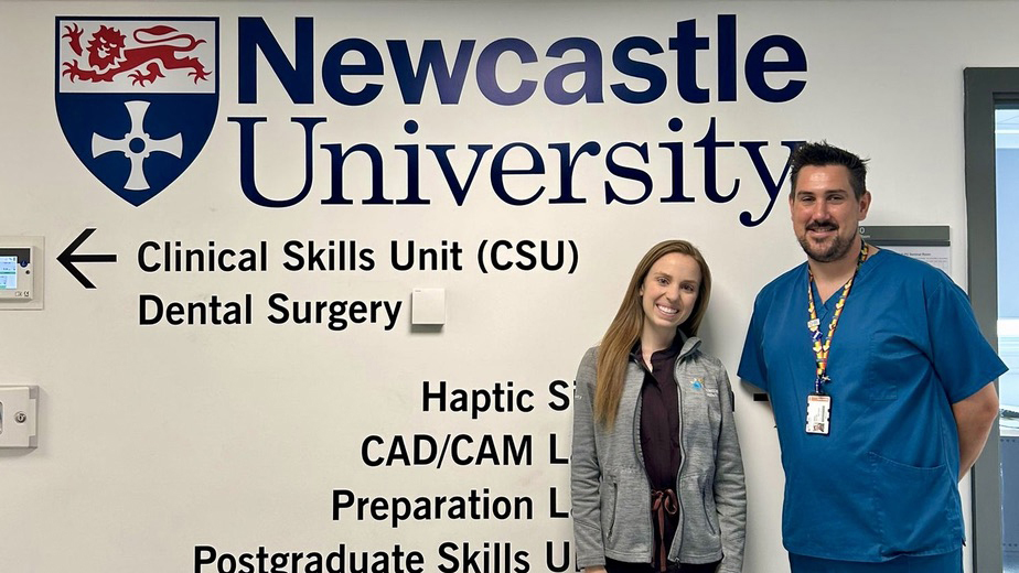 Amanda Swanson stands with a men dressed in blue in front of a wall that says Newcastle University