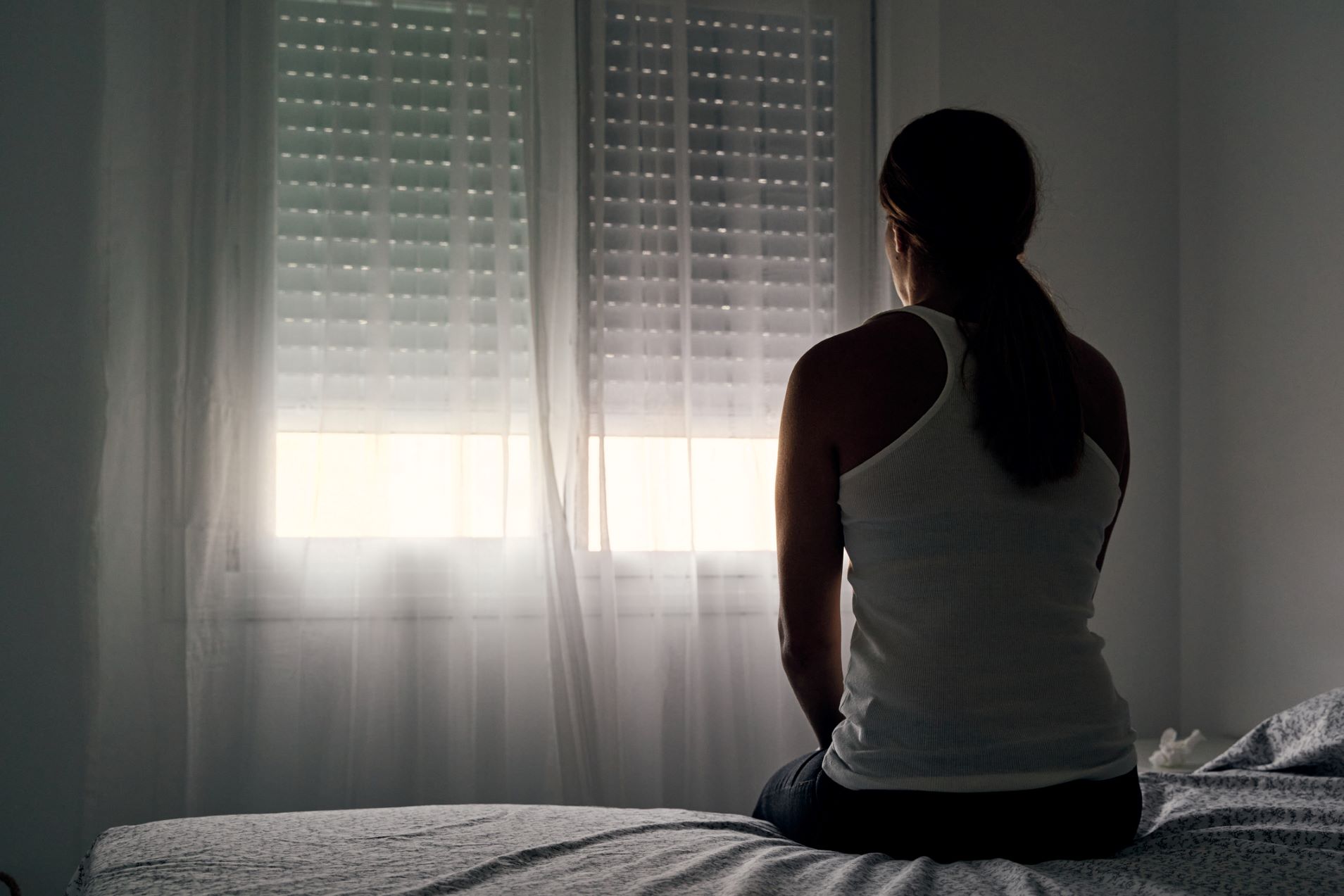 A woman sits on a bed looking toward a window