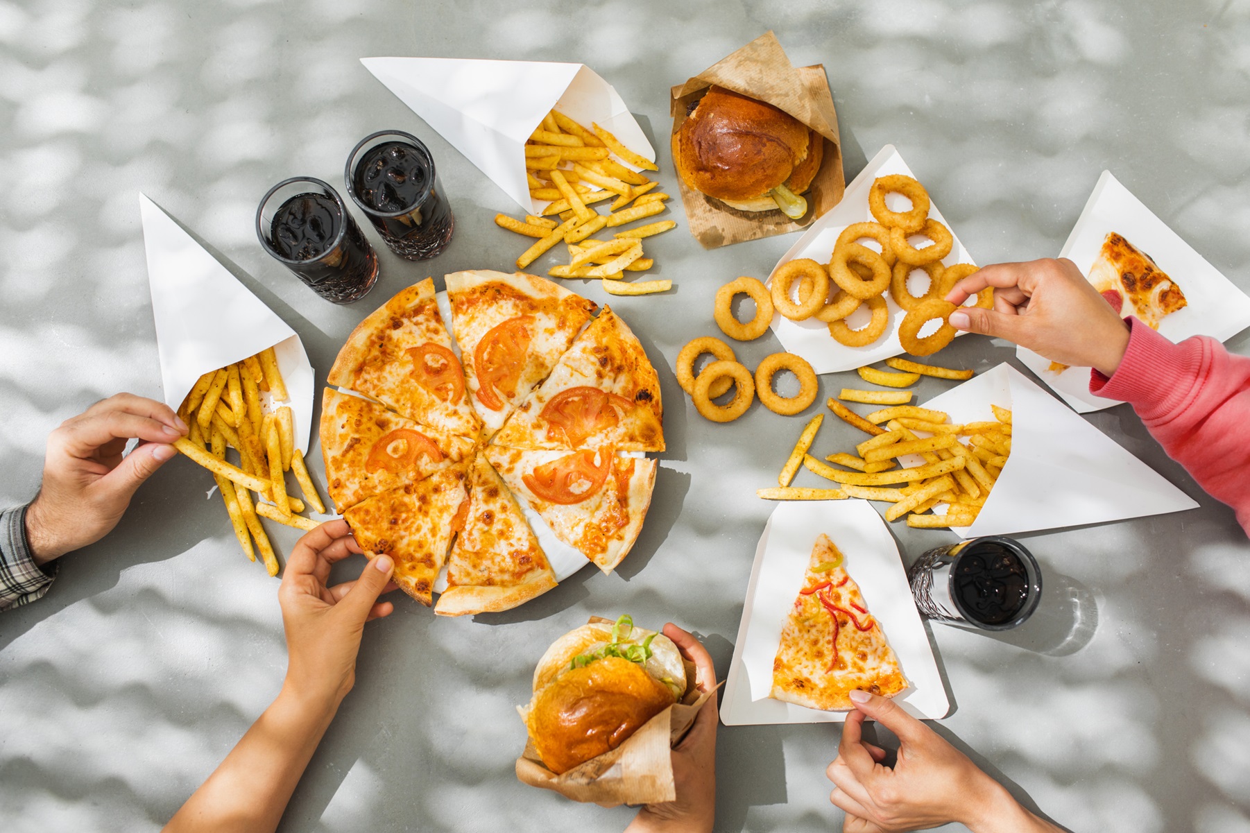 Hands reaching toward a table of foods including pizza, onion rings and burgers