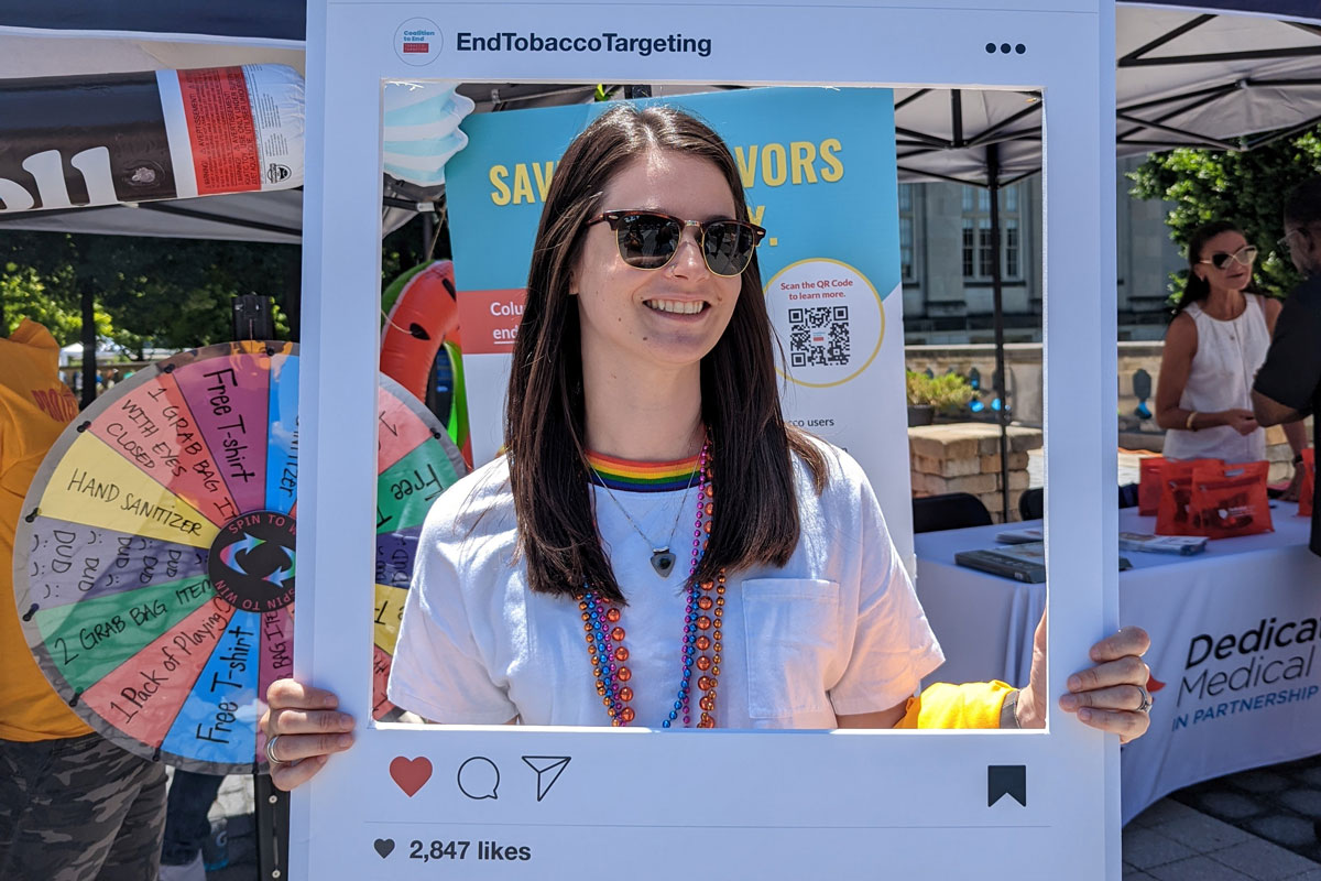 Young woman wearing sunglasses holds an Instagram-style sign at an outdoor health event
