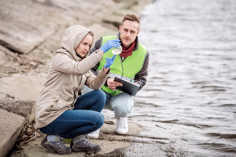 Two scientists crouch near a shoreline while examining water in a beaker