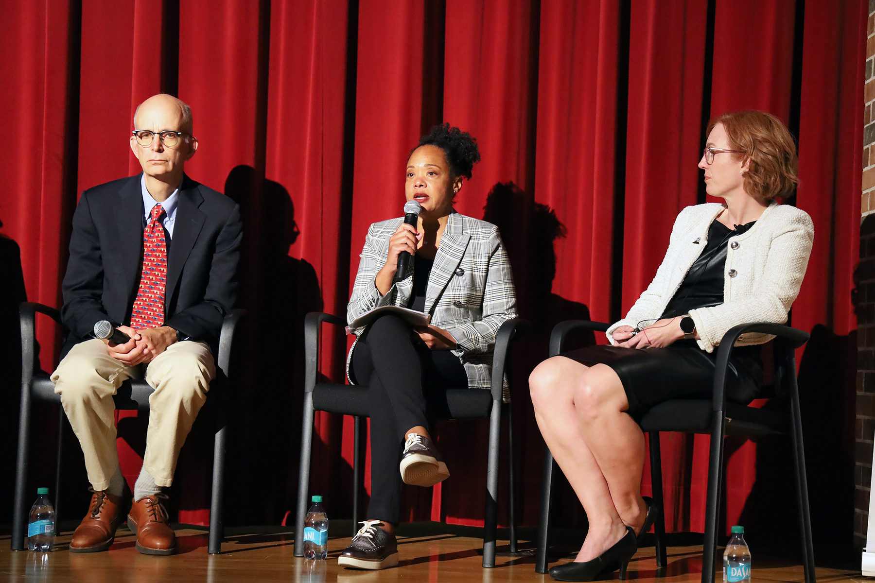 three panelists sit on stage while the person in the center speaks into a microphone.