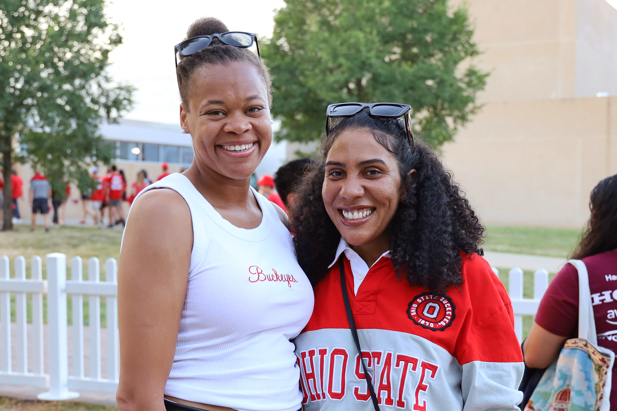 two women smile outdoors with a white fence in the background.