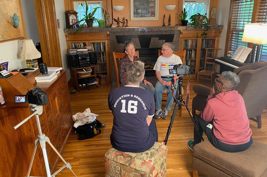 Four people sit in a living room. Two people are being interviewed on camera.