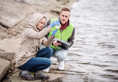 Two scientists crouch near a shoreline while examining water in a beaker 