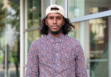 Sharmake standing in front of Cunz Hall wearing baseball hat backwards