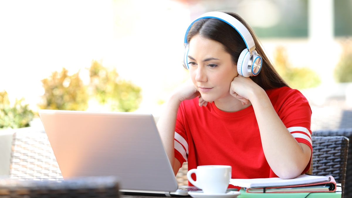 Woman wearing over-the-ear headphones uses computer and notebook to study at table outdoors