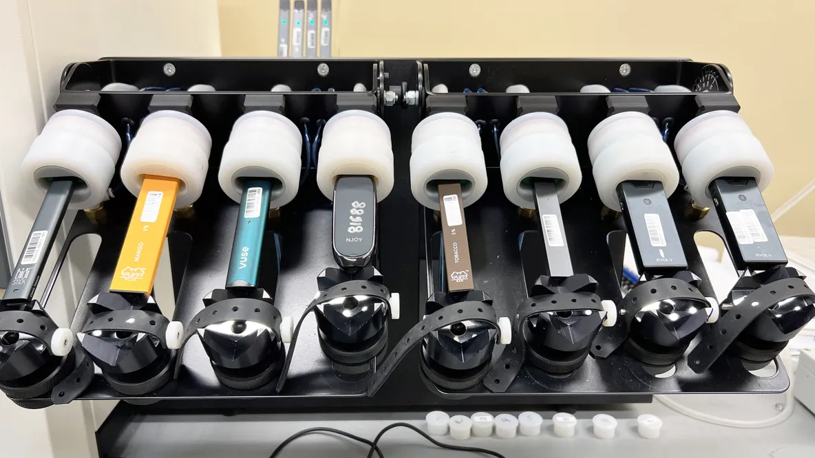  Eight e-cigarettes of different colors and sizes are plugged into white cylinders for emissions testing. 