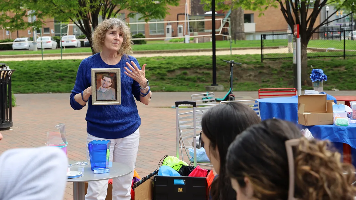 A woman dressed in blue holds a photo while addressing seated individuals. They are outside. 
