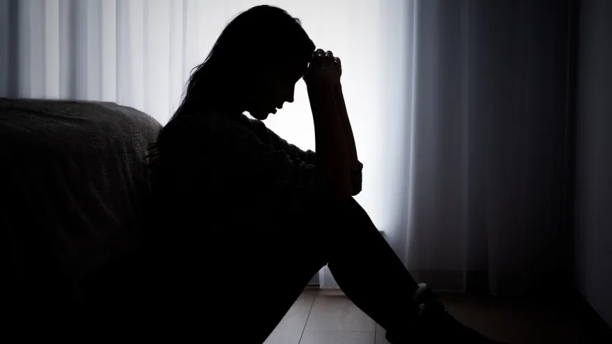 A woman in sillhouette, with her hands pulled to her forehead, sitting on a floor in front of a white curtain.