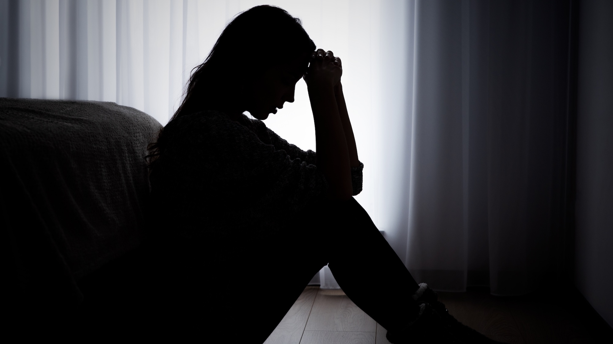 A woman in sillhouette, with her hands pulled to her forehead, sitting on a floor in frotn of a white curtain.