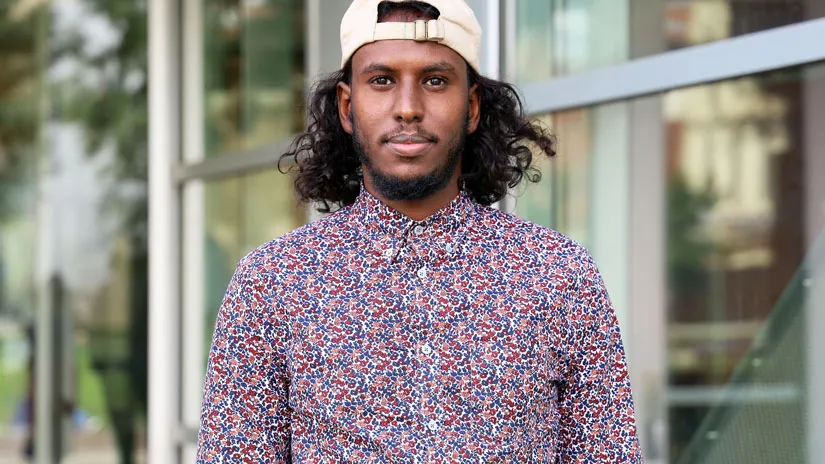 Sharmake standing in front of Cunz Hall wearing baseball hat backwards