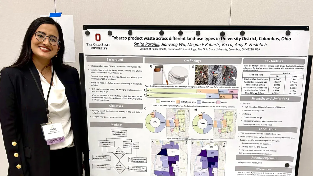 A smiling person in business attire stands next to an academic research poster with photos and graphics. 