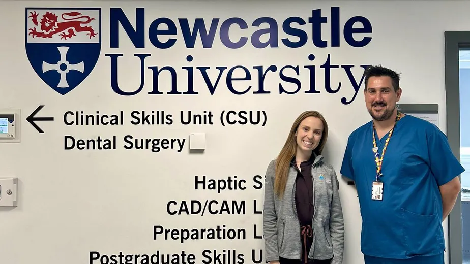 Amanda Swanson stands with a men dressed in blue in front of a wall that says Newcastle University