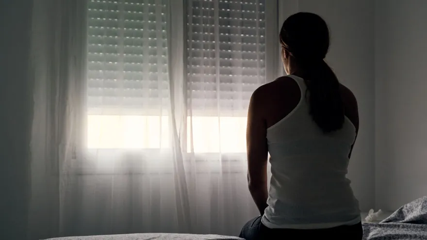 A woman sits on a bed looking toward a window