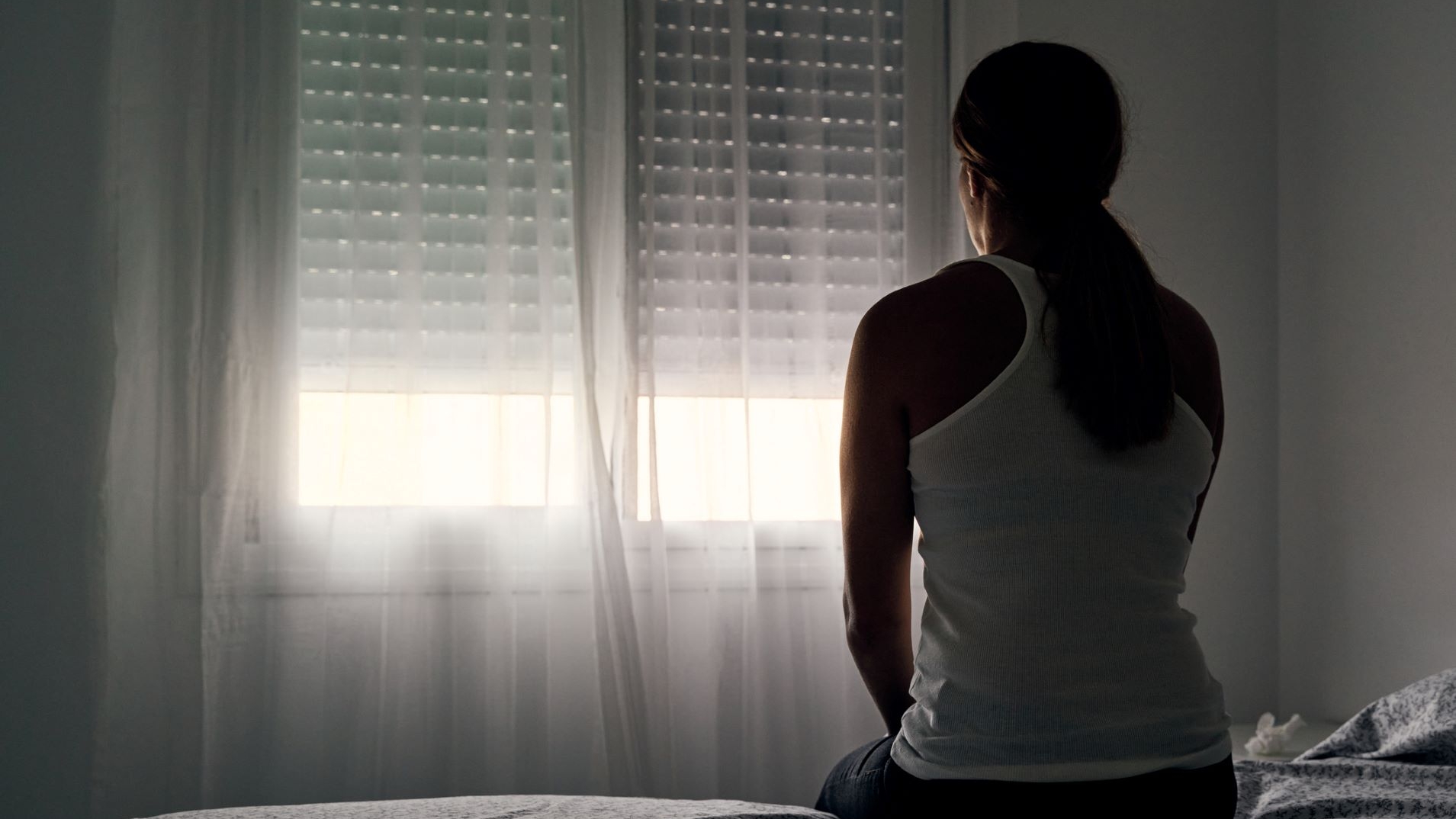 A woman sits on a bed looking toward a window