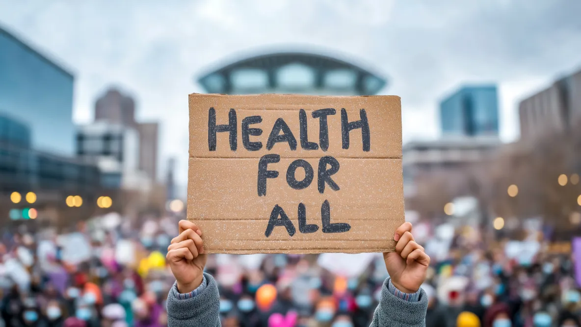 Hands hold up a sign that says "Health for all" with lots of people around.