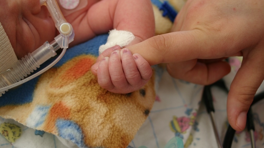 A hospitalized infant with a breathing apparatus on its face holds onto the finger of an adult
