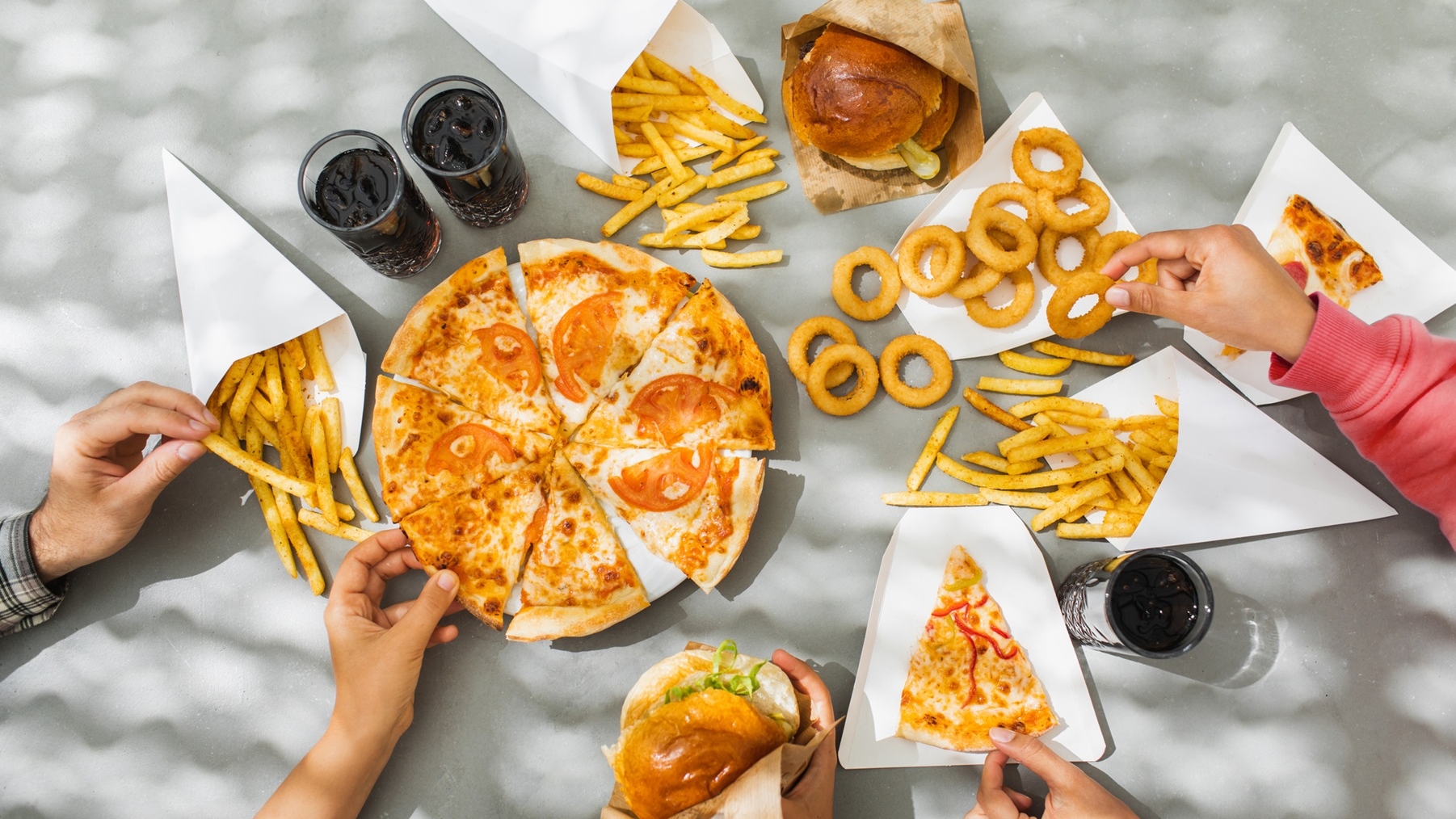 Hands reaching toward a table of foods including pizza, onion rings and burgers