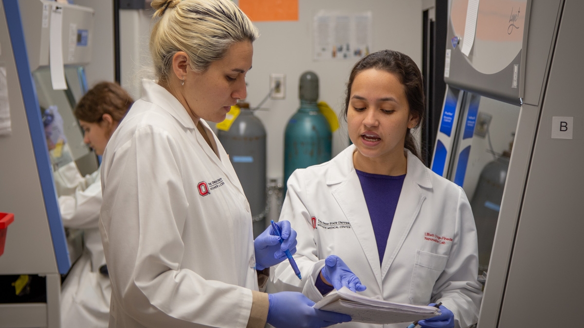 Two women in lab coats talk about a research report they are holding