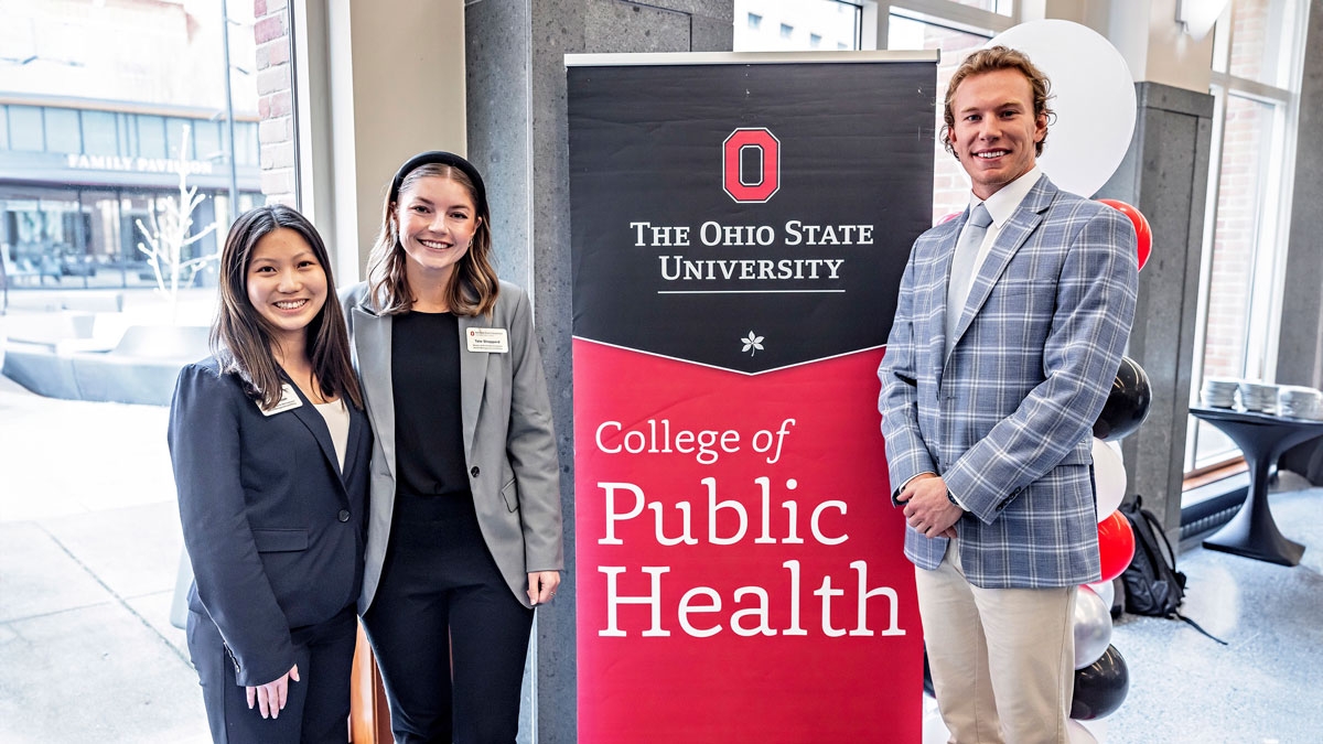 Three MHA students stand in a lobby with banner sign