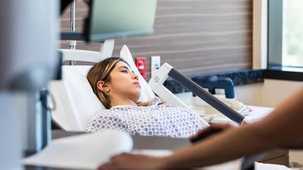 A young woman in a hospital bed with a health care professional's arm in the foreground