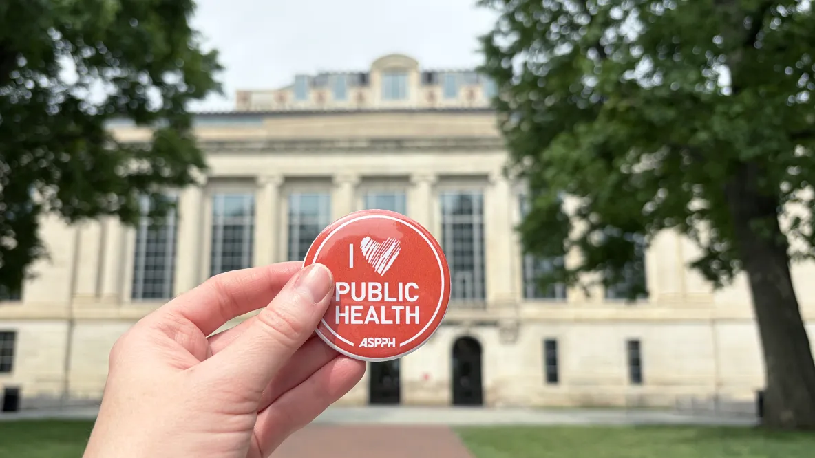 A hand holds a button that reads "I heart public health" and "ASPPH" in front of Thompson Library.