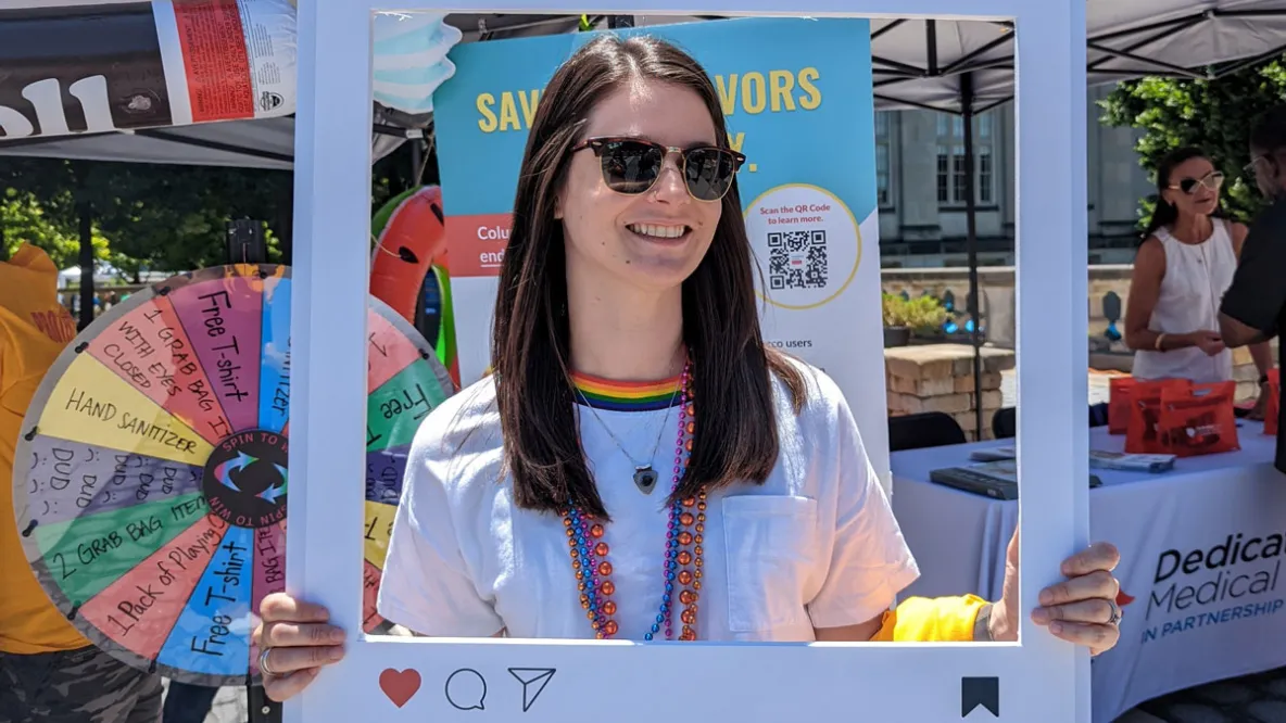 Young woman wearing sunglasses holds an Instagram-style sign at an outdoor health event