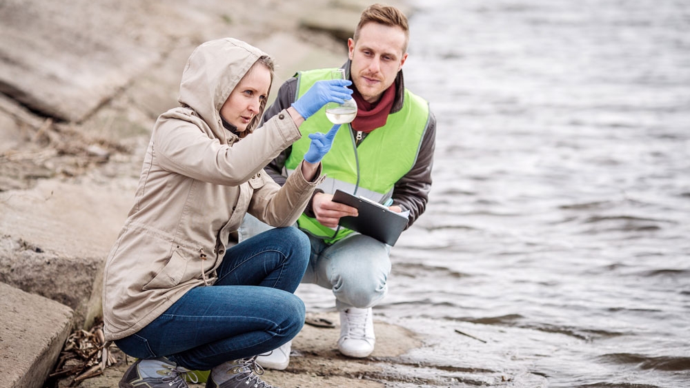 Two scientists crouch near a shoreline while examining water in a beaker