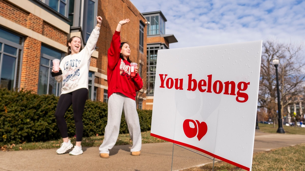 Two young women waving and walking by a sign that says "You belong"