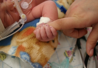 A hospitalized infant with a breathing apparatus on its face holds onto the finger of an adult